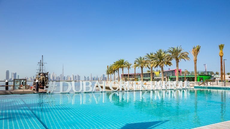 Dubai Creek Harbour sign by turquoise water infinity pool with rows of palm trees, Black Pearl Pirate Ship and Dubai Downtown skyline in the background, panoramic view.