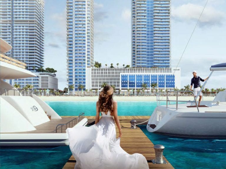 lady walking in white dress on the wharf between yachts in front of emaar beachfront residential buildings in the distance